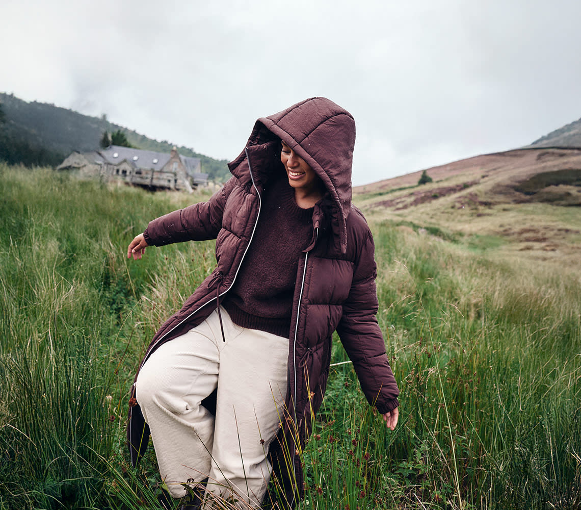 A woman in a brown coat and cream pants stands amidst tall grass, surrounded by a natural landscape.
